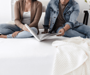 Man and woman sitting on a bed reading a book together they are sharing - showing chronic illness and relationships. 
