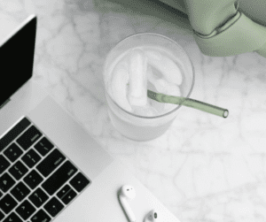 Image of a glass of water with ice cubes and a green straw sticking out of the cup beside a laptop. Showing the importance of staying hydrated while working to help reduce dehydration and migraines. 
