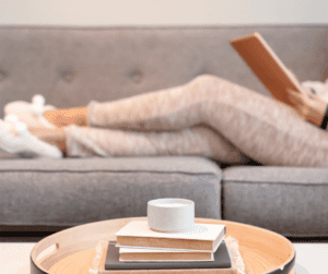 Image of woman laying on a grey couch. There is a pile of book at the front of the image. 