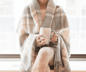 Image of a woman sitting in a blanket with a mug. Showing staying comfortable with fibromyalgia and learning more about what are the symptoms of fibro fog. 